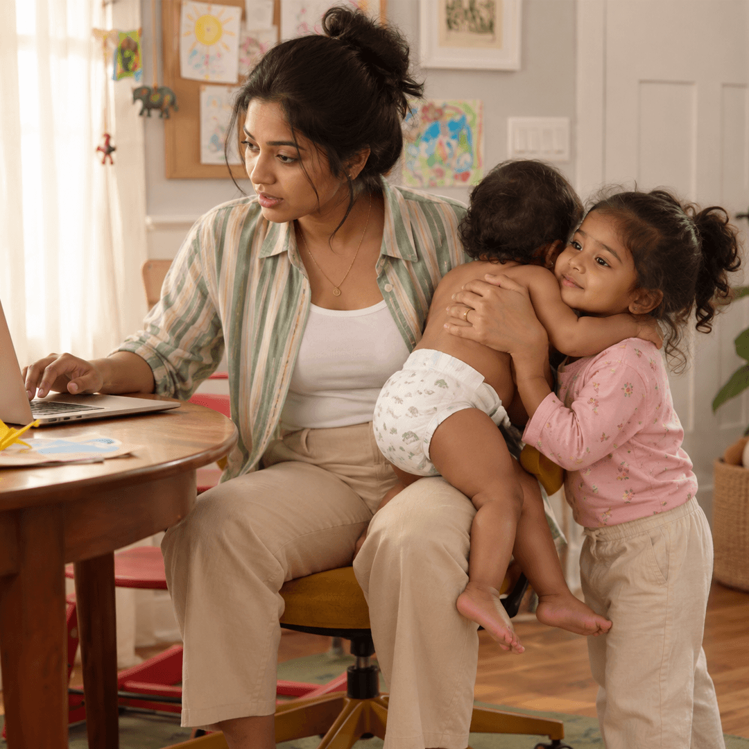 Mother working at a laptop while a toddler hugs the baby beside her